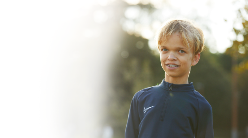 Un garçon aux cheveux blonds, atteint d'achondroplasie, qui porte un appareil dentaire et un maillot de course.