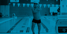 a boy with Achondroplasia posing in front of a swimming pool with a swim cap, bathing suit and goggles