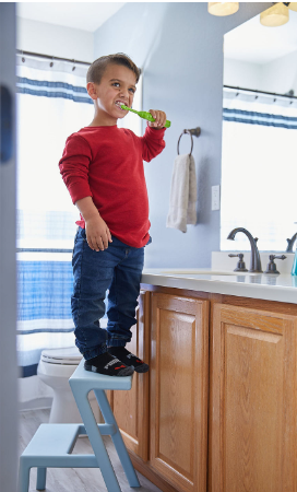 a boy with Achondroplasia standing on a stool brushing his teeth in the bathroom